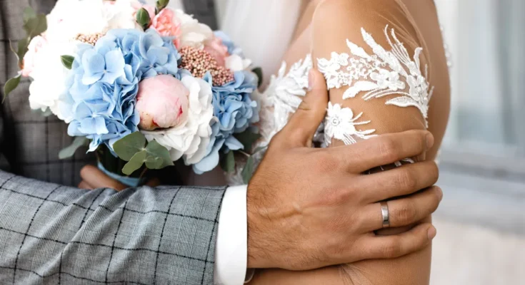 A groom gently holds his bride, their hands wrapped around a bouquet of pastel flowers | Source: Freepik